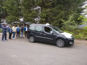 Attendees standing around a taxi listening to Colin Jones explaining what they will help a wheelchair customer into the back of the cab.